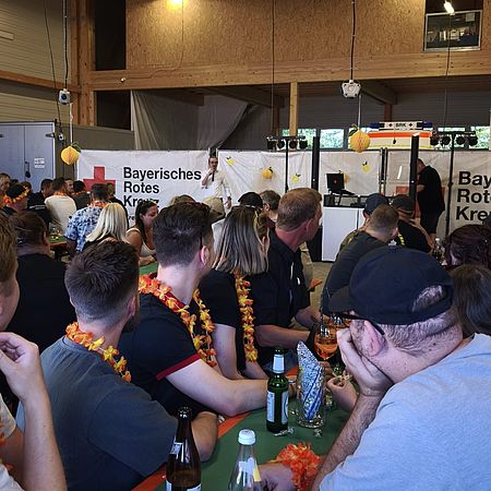 Sommerfest Dieses Bild zeigt mehrere Personen mit einer schwarz/rot/goldenen Blumengirlande um den Hals an Tischen sitzen. Im Hintergrund befinden sich weiße Banner, auf denen das BRK-Logo abgebildet ist.