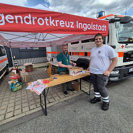 JRK Dieses Bild zeigt zwei Einsatzkräfte des Jugendrotkreuzes, die unter einem Pavillon hinter großen Tischen stehen. Auf den Tischen befinden sich Informationsmaterial und Werbegeschenke. Daneben befindet sich ein BRK-Einsatzfahrzeug.