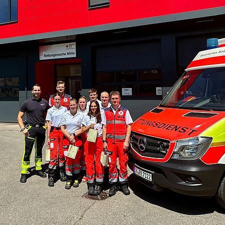 Praxisanleitertraining Dieses Bild zeigt mehrere Einsatzkräfte in Dienskleidung, die in der Rettungswache neben einem Rettungswagen stehen und in die Kamera lächeln.