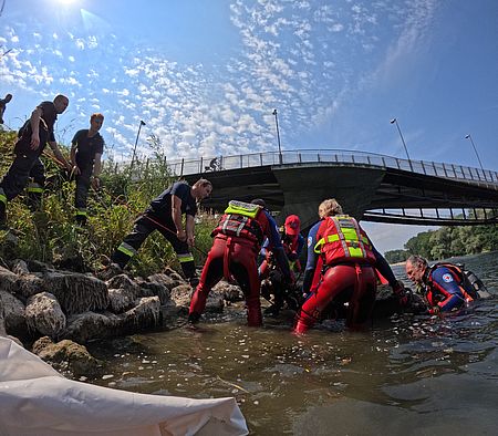 WW Bergung Dieses Bild zeigt mehrere Einsatzkräfte der Wasserwacht, die eine Person aus dem Wasser bergen. Im Hintergrund ist eine Brücke zu sehen.